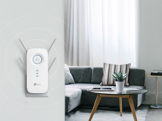TP-Link RE650 WiFi-repeater in een woonkamer, aangesloten op de muur, naast een grijze bank en salontafel met plant.