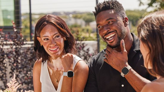 Een lachend stel buiten, beiden dragen Fitbit Sense 2 smartwatches met lichtblauwe bandjes.
