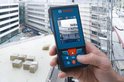 Hand die een Bosch laser afstandsmeter vasthoudt met een stadsgezicht en bouwplaats op de achtergrond.