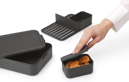 Hand pakt klein bakje met partjes sinaasappel uit een donkergrijze bento lunchbox. De lunchbox heeft een deksel en een scheidingswand.