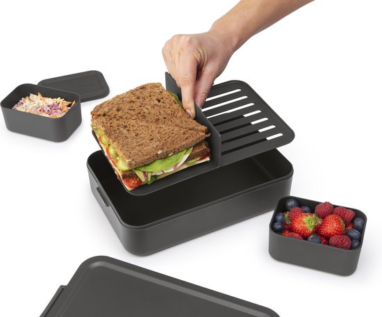 Hand legt een sandwich op een donkergrijze, gesegmenteerde lunchbox. Vruchten en salade ernaast.