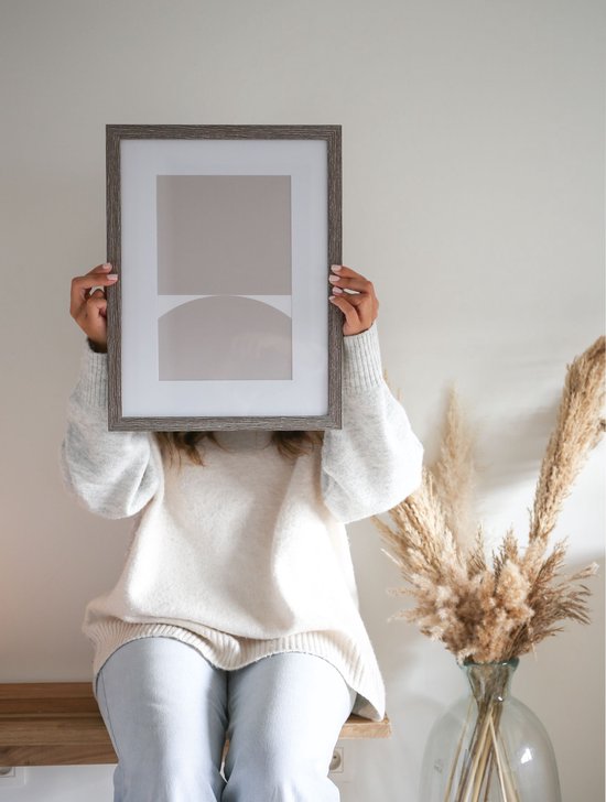 Femme en pull tricoté blanc tenant un cadre gris devant son visage, avec de l'herbe de pampa à côté.
