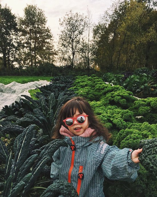 Meisje met zonnebril in groen regenpak met visgraatpatroon, staand tussen kale.