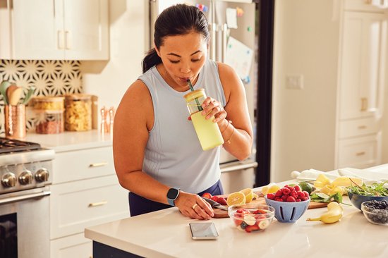Vrouw met zwarte Fitbit Versa 4 smartwatch in keuken bij bereiden van fruit.