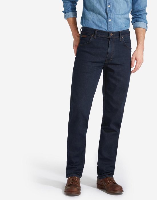 Model draagt een donkerblauwe Wrangler jeans met regular fit, gecombineerd met een spijkerhemd en bruine schoenen.