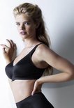 Blanke vrouw met blond haar, draagt zwarte beugel-bh met brede bandjes en V-hals, leunend tegen witte muur.