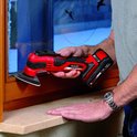 Een man met een grijze trui houdt een rode en zwarte multitool met accu vast en schuurt een houten kozijn.