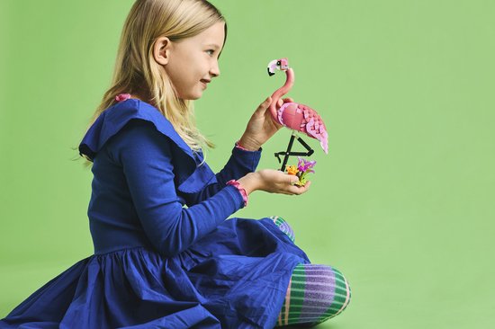 Een lachend meisje met blond haar, in een blauwe jurk, houdt een roze LEGO flamingo vast met bloemen.