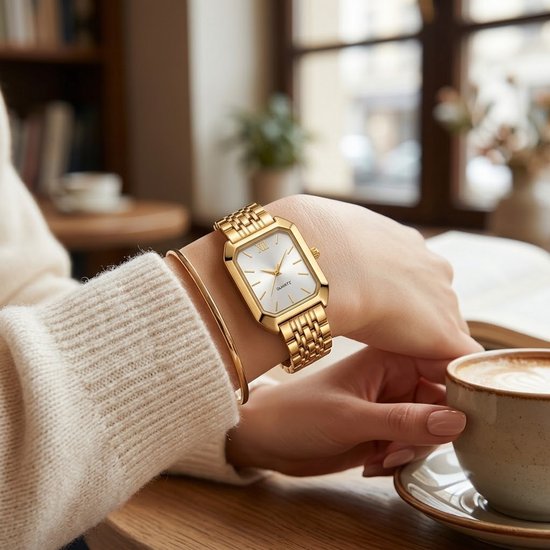 Gouden rechthoekig analoog horloge met witte wijzerplaat en Romeinse cijfers, gedragen om een pols, naast een kop koffie.