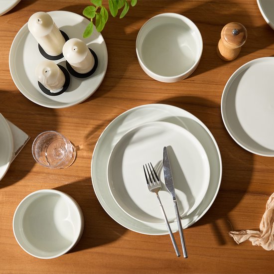 Bovenaanzicht van witte borden en kommen op een houten tafel, met kaarsen en een pepermolen.