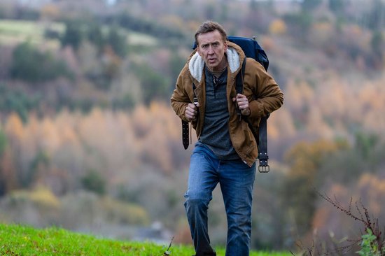 Nicolas Cage draagt een rode jas, grijs vest en spijkerbroek met een rugzak.