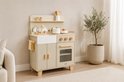 Beige houten speelkeuken met witte spoelbak, oven en kookplaat. Accessoires zoals potten, borden en bestek zijn aanwezig. Een plant staat ernaast.
