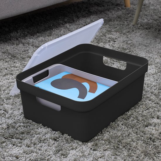 Zwarte opbergbox met handgrepen op een grijze vloer, met een transparante deksel ernaast. Binnenin liggen een dienblad en bruine objecten.