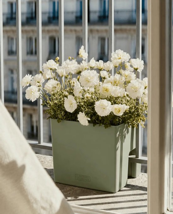Olijfgroene plantenbak vol witte bloemen, bevestigd aan een balkonhek met zilveren spijlen.