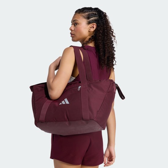 Vrouw draagt een bordeauxrode sporttas met zilverkleurig Adidas logo op de schouder.