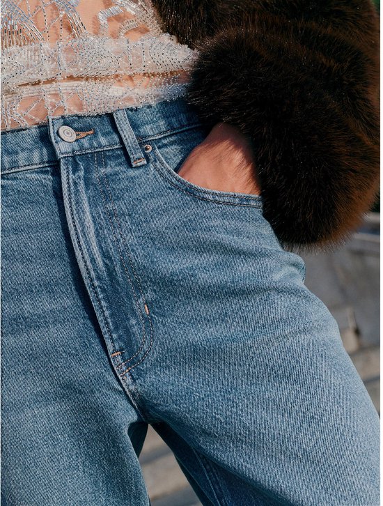 Model met een hand in de broekzak, draagt een spijkerbroek met hoge taille en een donkerbruine jas met bontkraag.