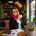 Een vrouw met bril draagt een rood kersenpitkussen om haar nek, drinkt koffie en werkt aan een laptop.