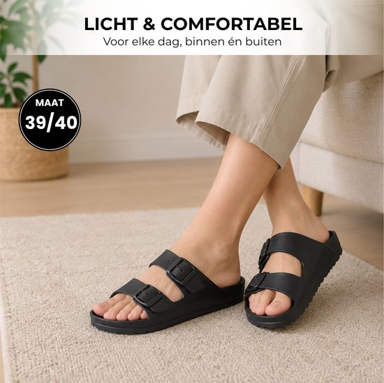 Voeten van een persoon die zwarte slippers met dubbele gespen dragen op een lichte vloer.
