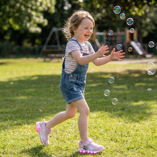Meisje in spijkerbroek met tuinbroek en gestreept shirt vangt bellen op grasveld met roze lichtgevende Frozen sneakers aan.