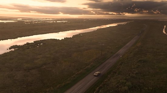 Vogelvlucht van een auto op een landweg die door drassige gebieden slingert bij zonsondergang.
