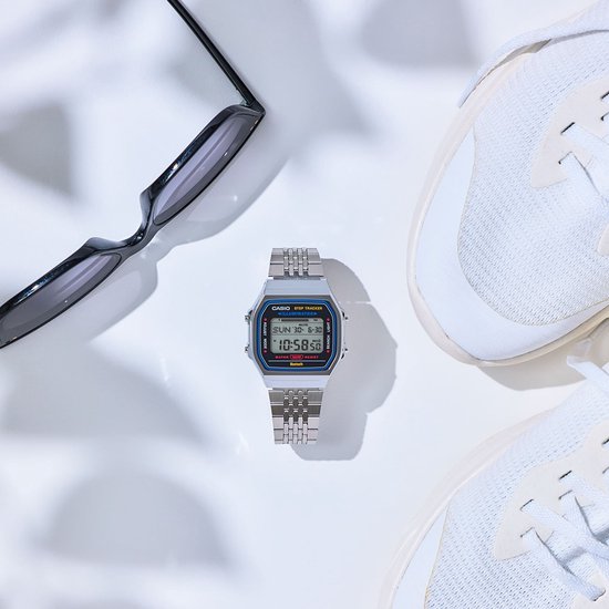 Stilleven met een zilverkleurig Casio horloge in het midden, geflankeerd door witte sneakers en een bril, tegen een witte achtergrond met schaduwen.