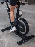 Deel van een persoon die trapt op een zwarte Fit Bike Race Magnetic Pro spinfiets met kleurrijke sokken en sportschoenen.