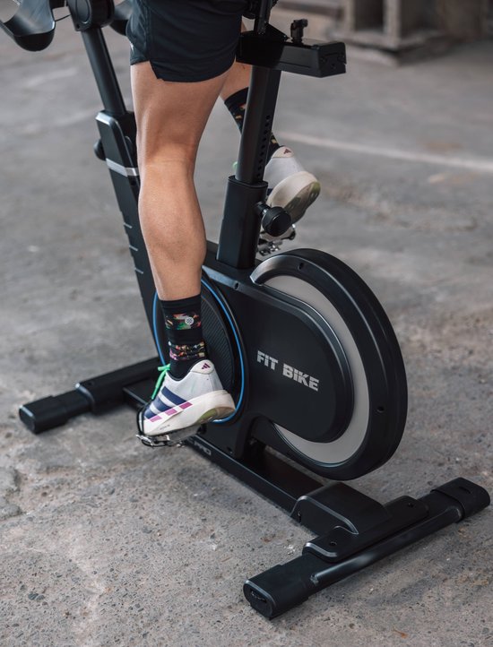 Deel van een persoon die trapt op een zwarte Fit Bike Race Magnetic Pro spinfiets met kleurrijke sokken en sportschoenen.