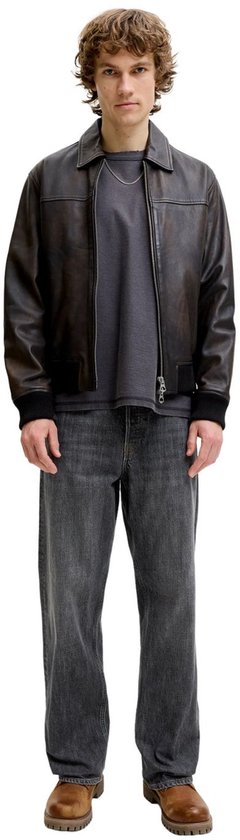 Model in een bruin leren jack, grijs T-shirt, grijze jeans en bruine schoenen.
