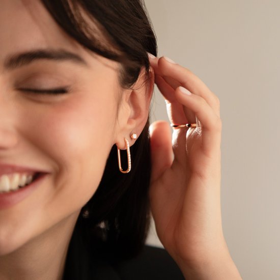 Een model draagt rechthoekige, roségouden oorbellen bezet met zirkonen. Een ring is zichtbaar op haar vinger.