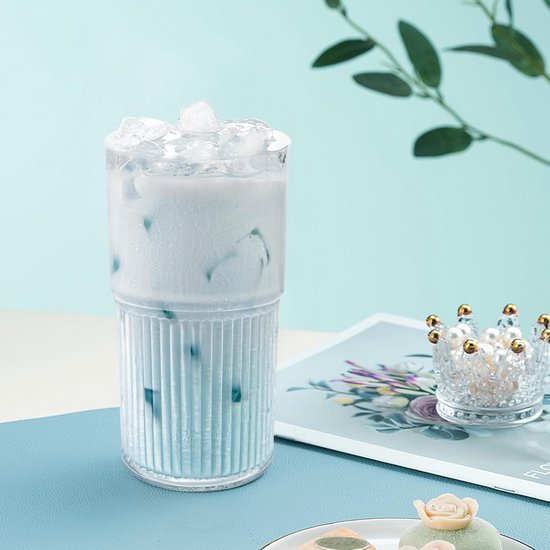 Geribbelde glazen beker gevuld met een lichtblauwe drank en ijs, op een blauwe ondergrond, met een takje en decoraties.