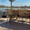 Beige tuinstoelen met ribbelpatroon en metalen poten aan een houten tafel op een houten terras aan het water.