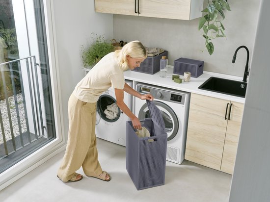 Een vrouw plaatst kleding in een antracietkleurige wasmand met deksel in een moderne wasruimte. De wasmand heeft een opengewerkt patroon.