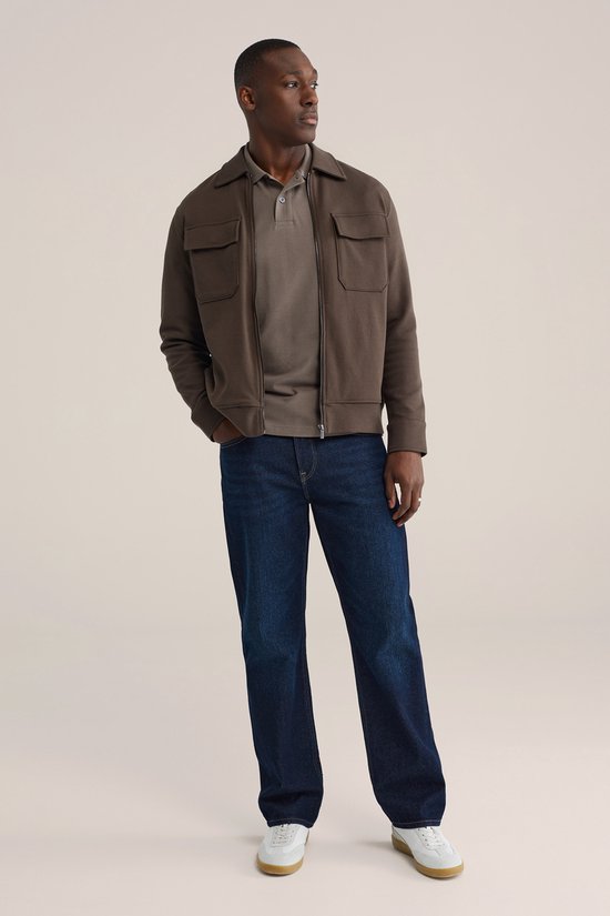 Man in bruine bomberjack met rits, grijs poloshirt en donkerblauwe jeans.