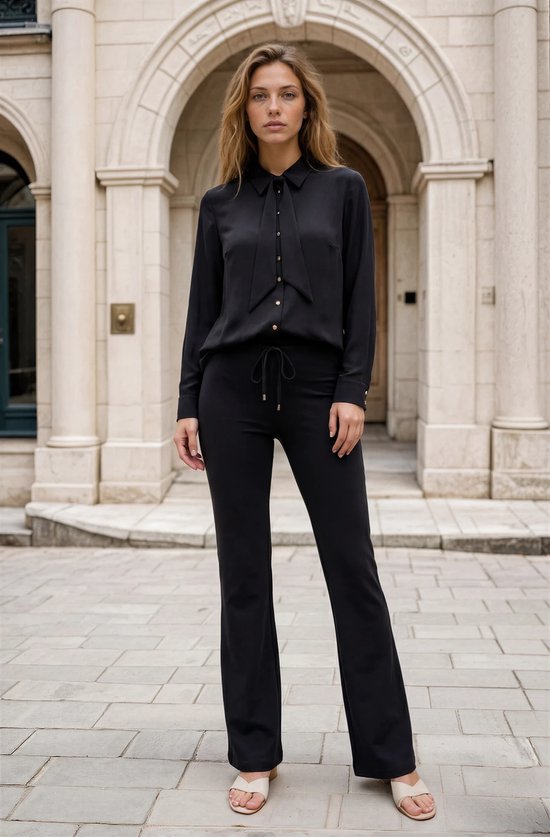 Model in zwarte broek met hoge taille, trekkoord en wijde pijpen. Draagt witte sandalen.