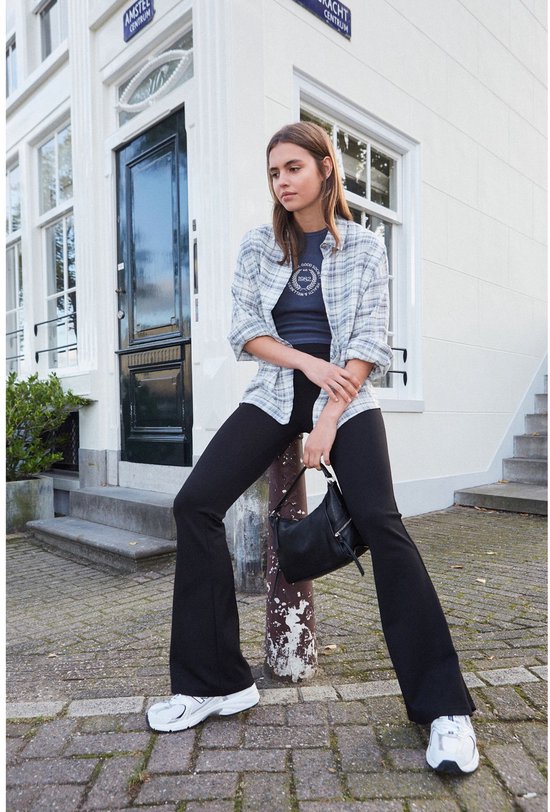 Vrouw zittend op een paal, draagt een zwarte wijduitlopende legging, grijs geruite blouse en witte sneakers.