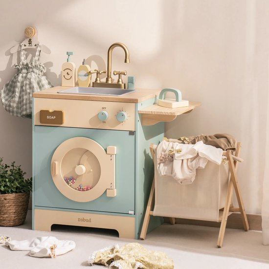 Houten speelgoed wasmachine in pastelblauw en beige, met gouden kraan, zeepdispenser en strijkplankje. Kleding hangt ernaast.