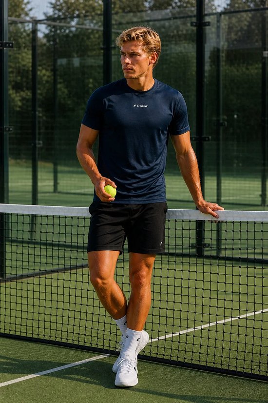 Man met een navy blauw RAQA sportshirt, zwarte shorts en witte schoenen, staand op een tennisbaan.