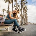 Een lachende vrouw in een turquoise sportoutfit zit op een houten bankje, draagt zwarte skeelers met witte wielen en houdt een koffiebeker vast.