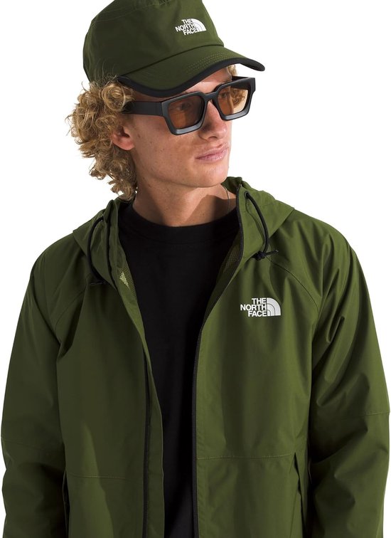 Close-up van man met groene The North Face outdoorjas met capuchon, zwart met rits, en zwarte top. Hij draagt een groene pet en een vierkante zonnebril.