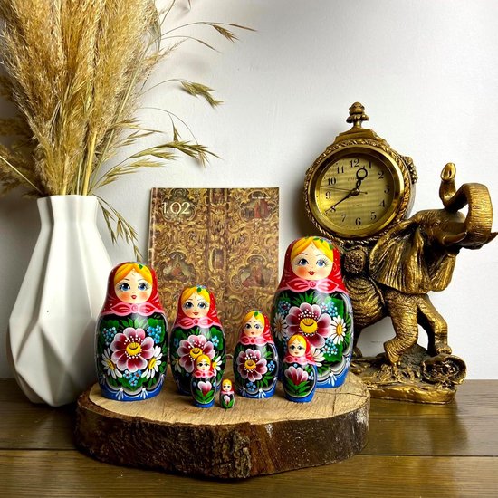 Zeven handgeschilderde Russische nestpoppen met bloemenpatronen, op een houten plak, naast een vaas en een gouden olifant.