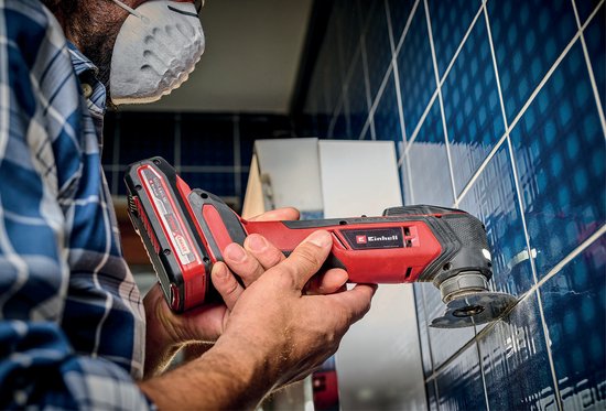 Man met rood-zwarte multitool en stofmasker snijdt in blauwe tegels.