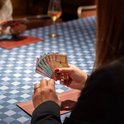 Een vrouw speelt een kaartspel aan een tafel met een blauw-wit geblokt tafelkleed. Ze houdt kaarten vast met afbeeldingen van wijn.