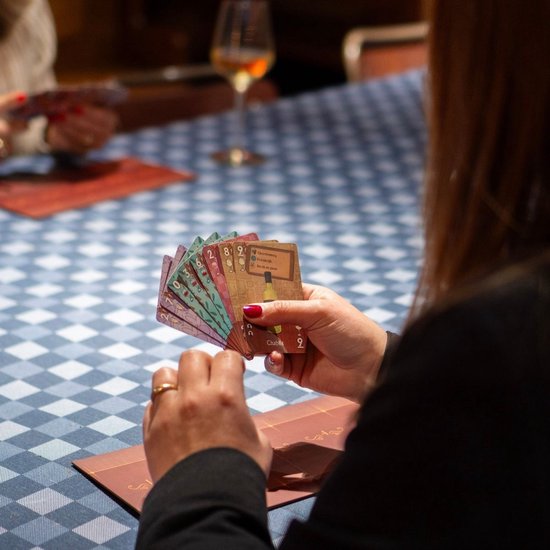 Een vrouw speelt een kaartspel aan een tafel met een blauw-wit geblokt tafelkleed. Ze houdt kaarten vast met afbeeldingen van wijn.
