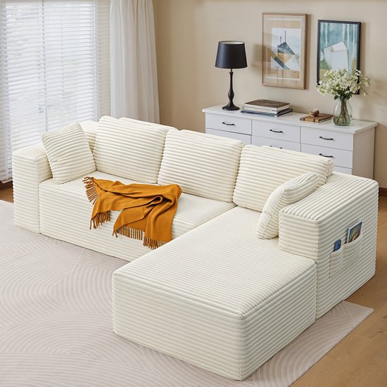 Beige corduroy L-vormige bank met kussens en een oranje plaid, met zijvakken met telefoon en pasjes.