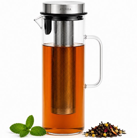 Carafe en verre avec infuseur en acier inoxydable, remplie de thé ambré. De la menthe et des feuilles de thé sont placées à côté de la carafe.