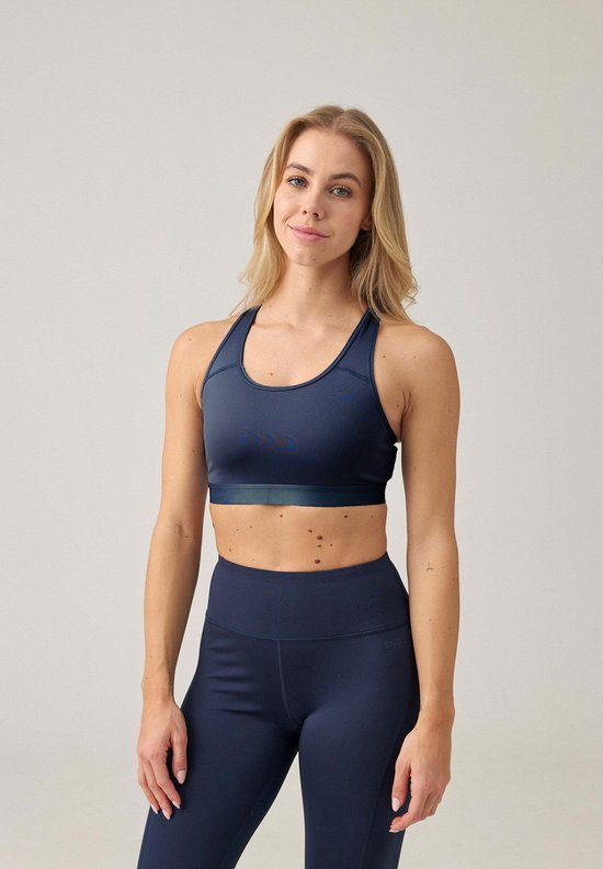Vrouw met blonde haren, draagt een donkerblauwe sportbeha met racerback en een bijpassende legging. Ze kijkt recht naar de camera met een glimlach.