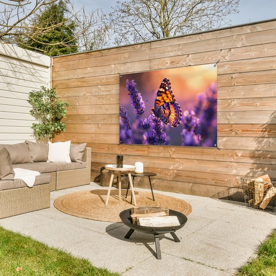 Een vlinder met oranje, zwart en paarse vleugels zit op paarse lavendel, in een tuin met rotan meubels.