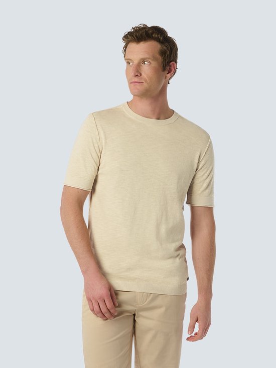Homme en t-shirt beige à manches courtes, debout, mains dans les poches.