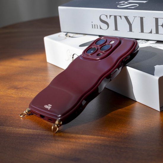 Glanzend bordeauxrood wavy telefoonhoesje voor iPhone 12 Pro Max, liggend op een tafel met boeken.