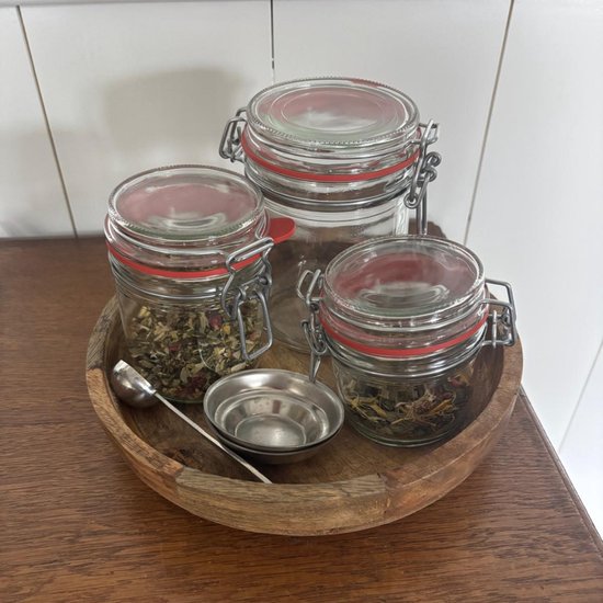 Houten schaal met drie glazen weckpotten van verschillende groottes gevuld met kruiden, inclusief een klein metalen schepje.